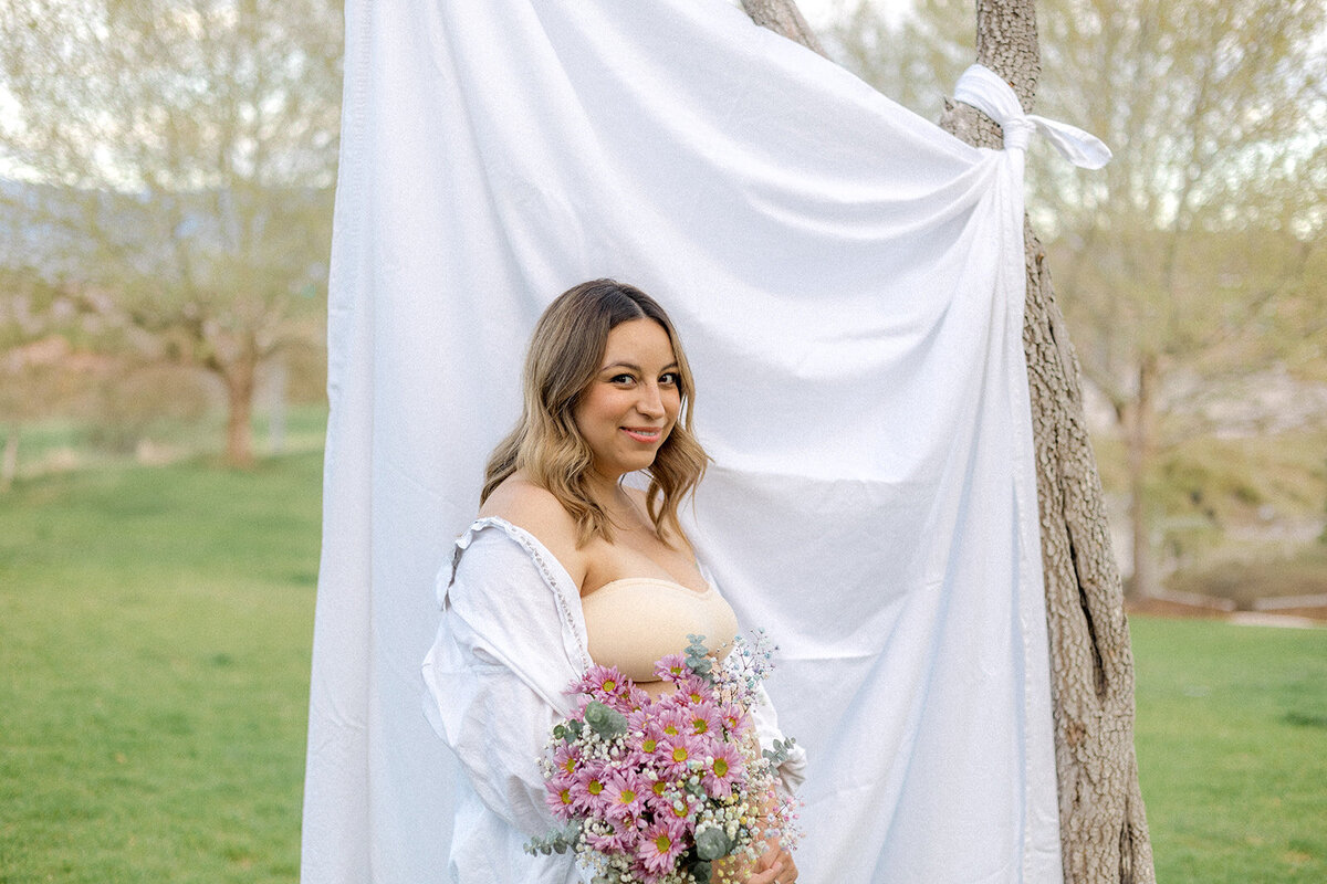 A pregnant woman stands in front of a sheet outdoors while holding flowers