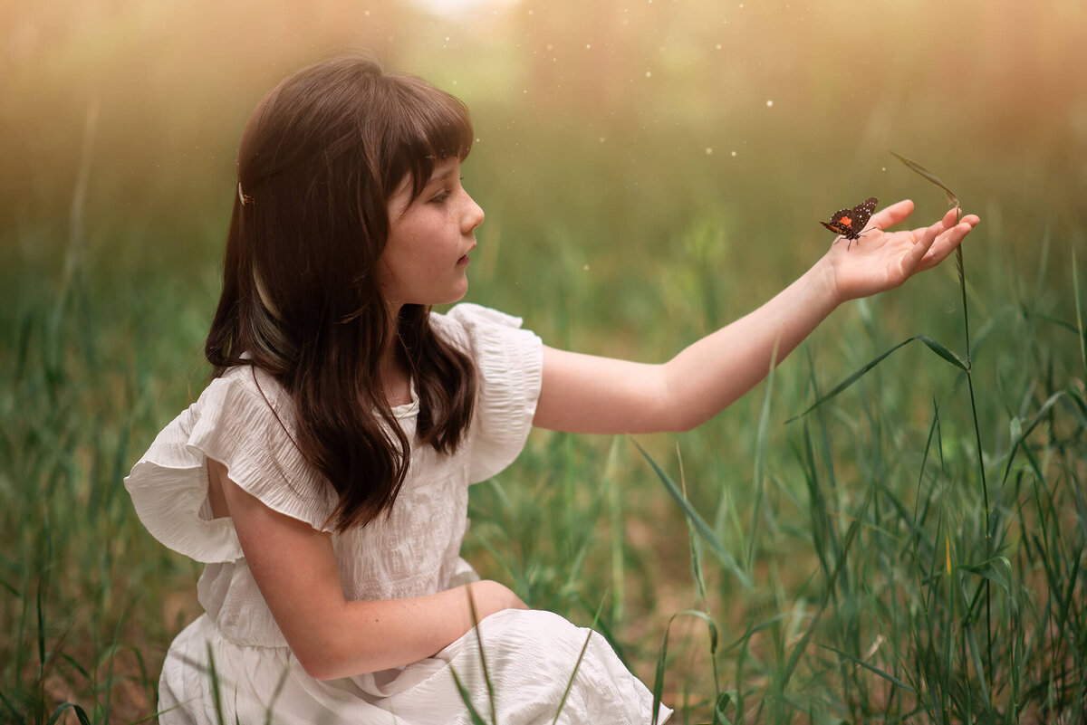 Springtime in Kamloops features a little girl with brown hair crouched in the grass holding out her arm with a butterfly on it. Taken by Brittany Danielle Photography.