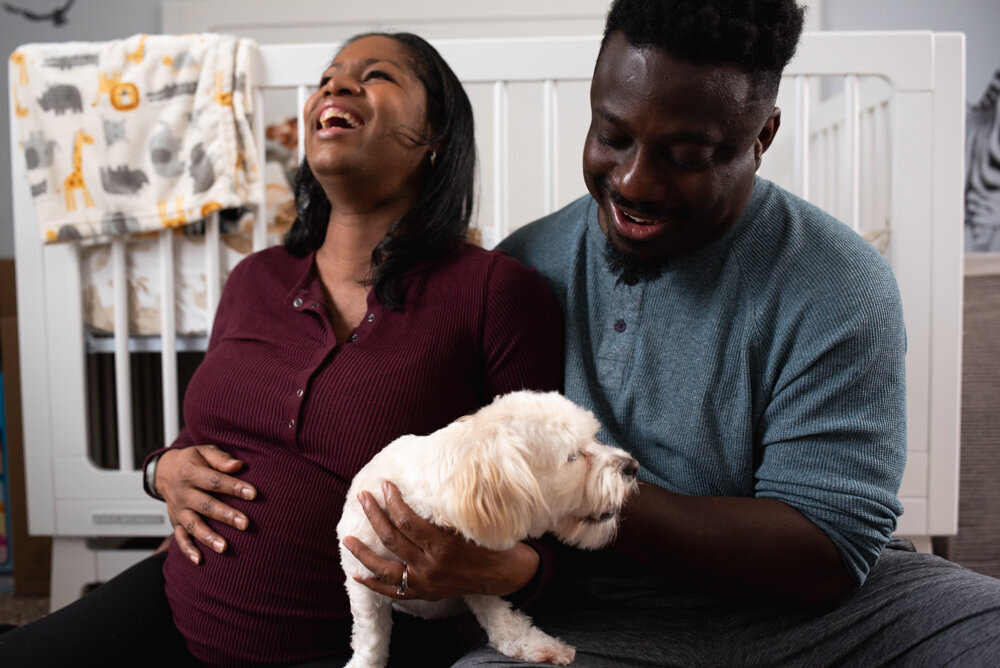 Family snuggled with their dog, laughing at the dog's antics while sitting on the floor of their baby-to-be's nursery for in home maternity session