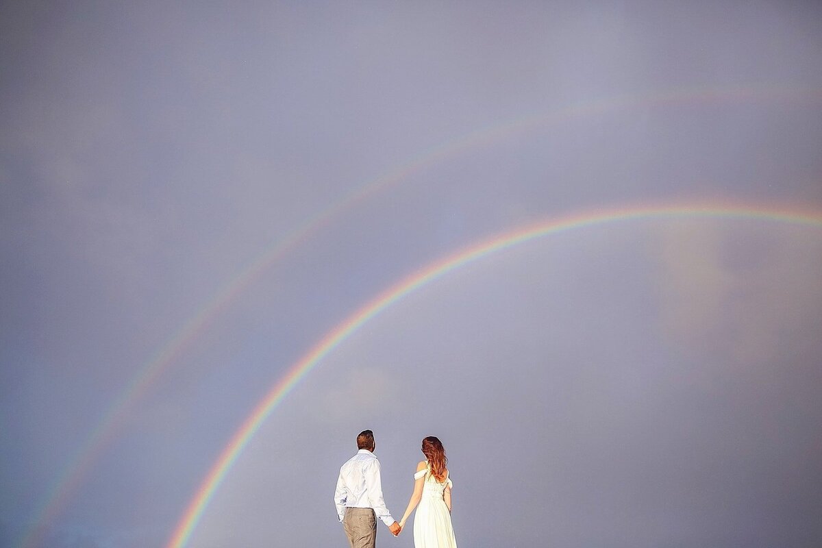 Woman wearing white dress holds her husband's hand as a double rainbow appears above them. They are sailing on a private yacht on Maui.
