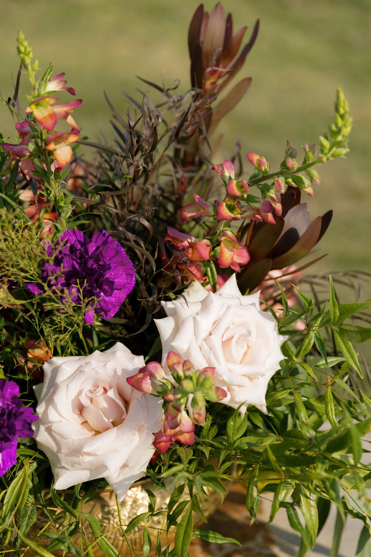 Romantic wedding floral arrangement with creamy roses, purple blooms, peach snapdragons, and rich foliage placed in a gold vessel for an elegant garden celebration.