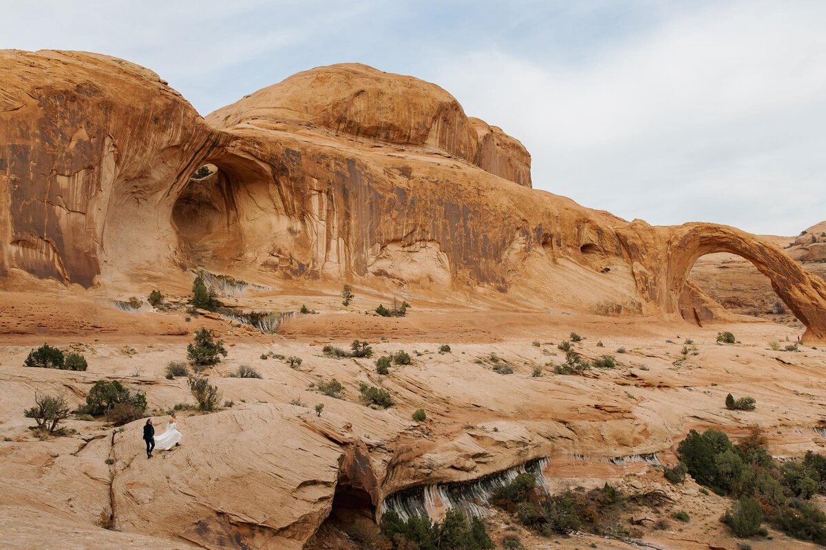 A groom helps carry his brides train as they walk towards Corona Arch for their elopement ceremony. 