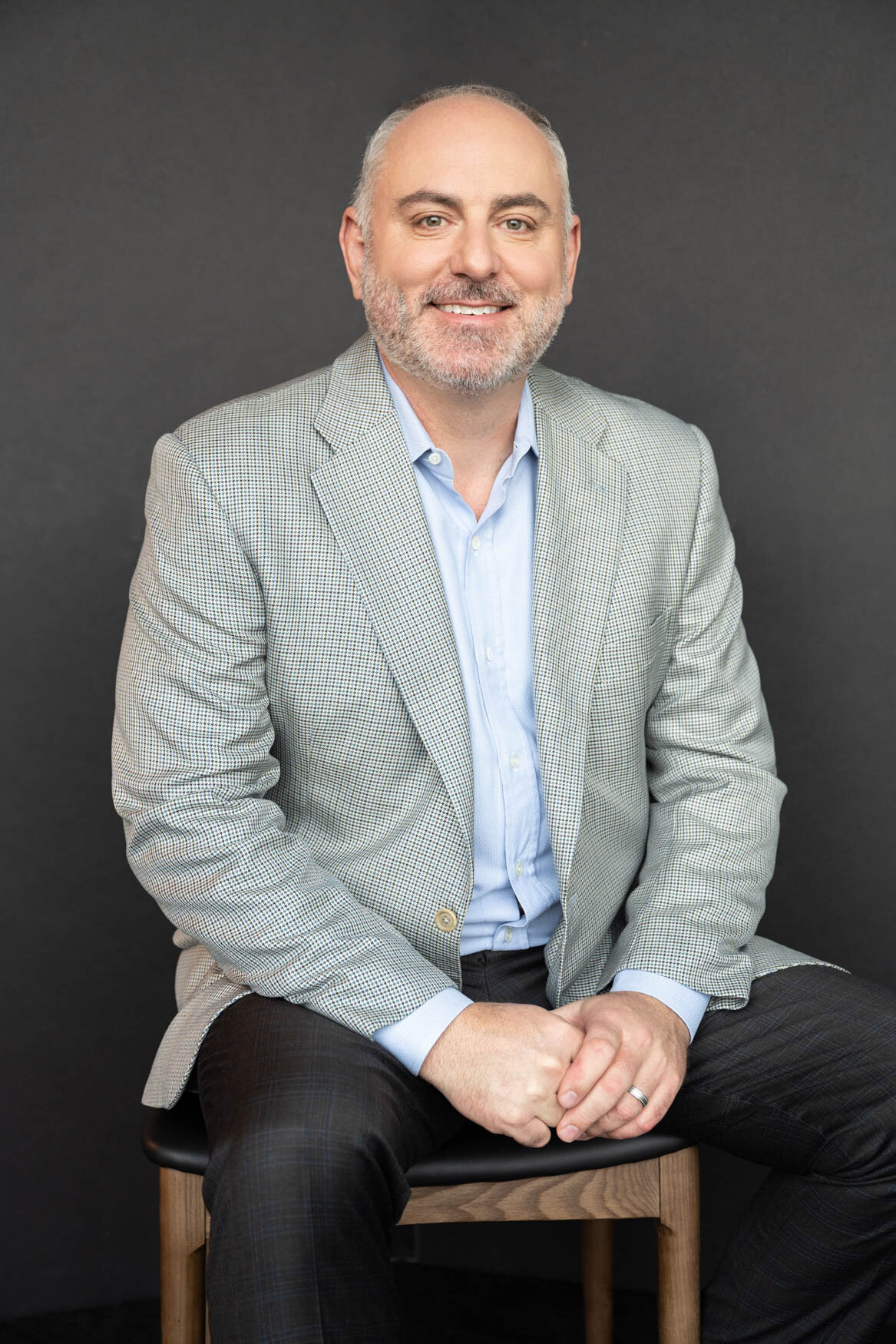 A portrait of a man seated  on a chair with his hands touching each other. He is wearing a grey suit jacket, black pants and is smiling to the camera