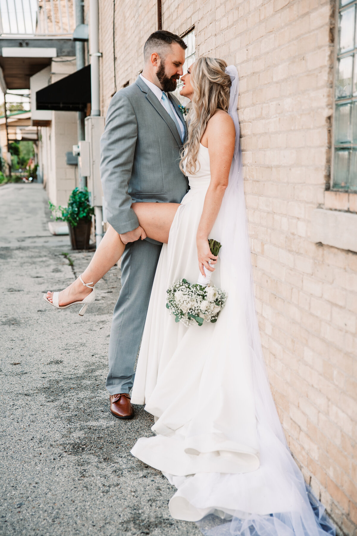 A groom holds his bride's leg up as they lean against a brick wall for a kiss