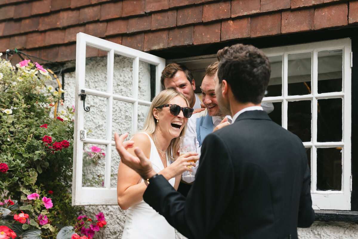 A bride in sunglasses laughs with three men through a window. She holds a drink and wears a white dress. The men are dressed in formal attire. Colorful flowers are visible in the foreground.