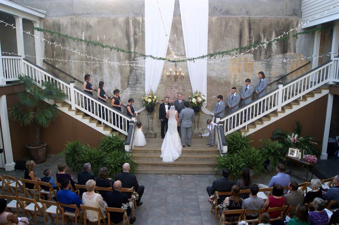 A wedding ceremony takes place outdoors between two staircases. The bride and groom stand at the altar with their wedding party flanking them on both sides, while guests watch from rows of wooden chairs.