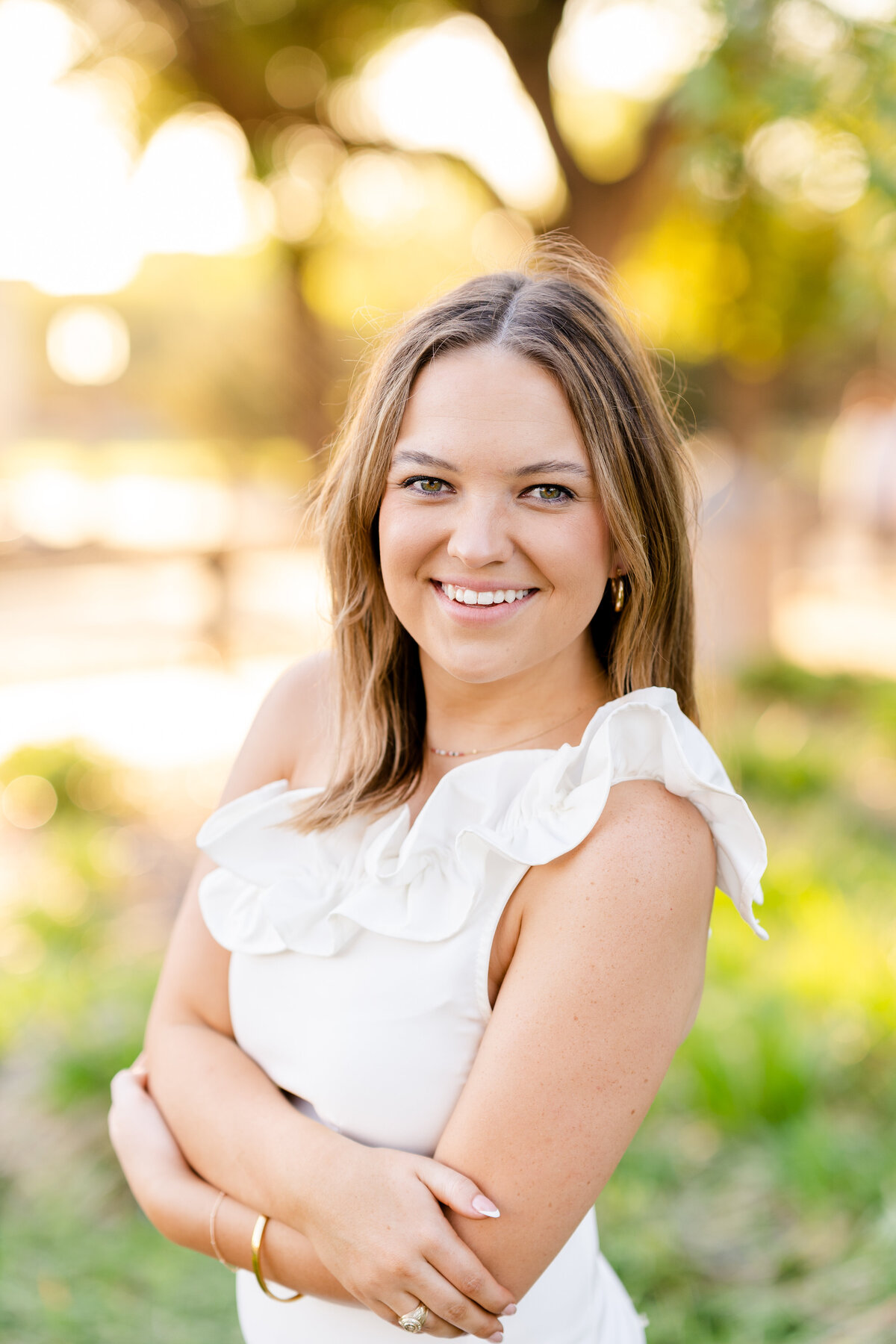 Texas A&M senior girl crossing arms and smiling while wearing white dress in glowy nature at sunset in Military Plaza