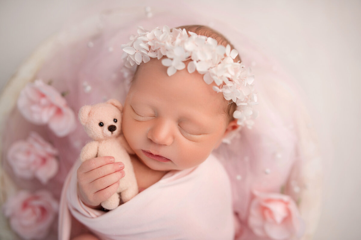 newborn baby girl wearing pink floral headband sleeping and holding teddy bear in white bowl surrounded by flowers