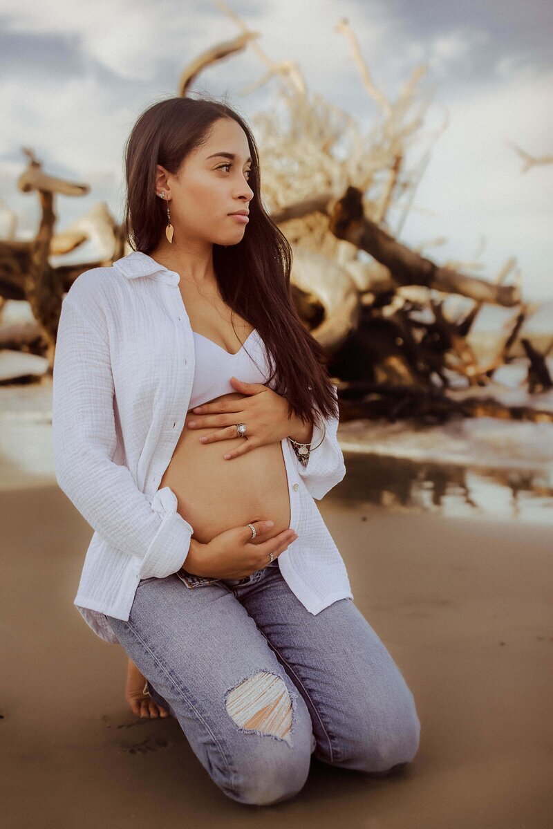 A pregnant woman kneels on a beach, wearing a white shirt and jeans, holding her belly with one hand. Driftwood and the ocean are in the background.