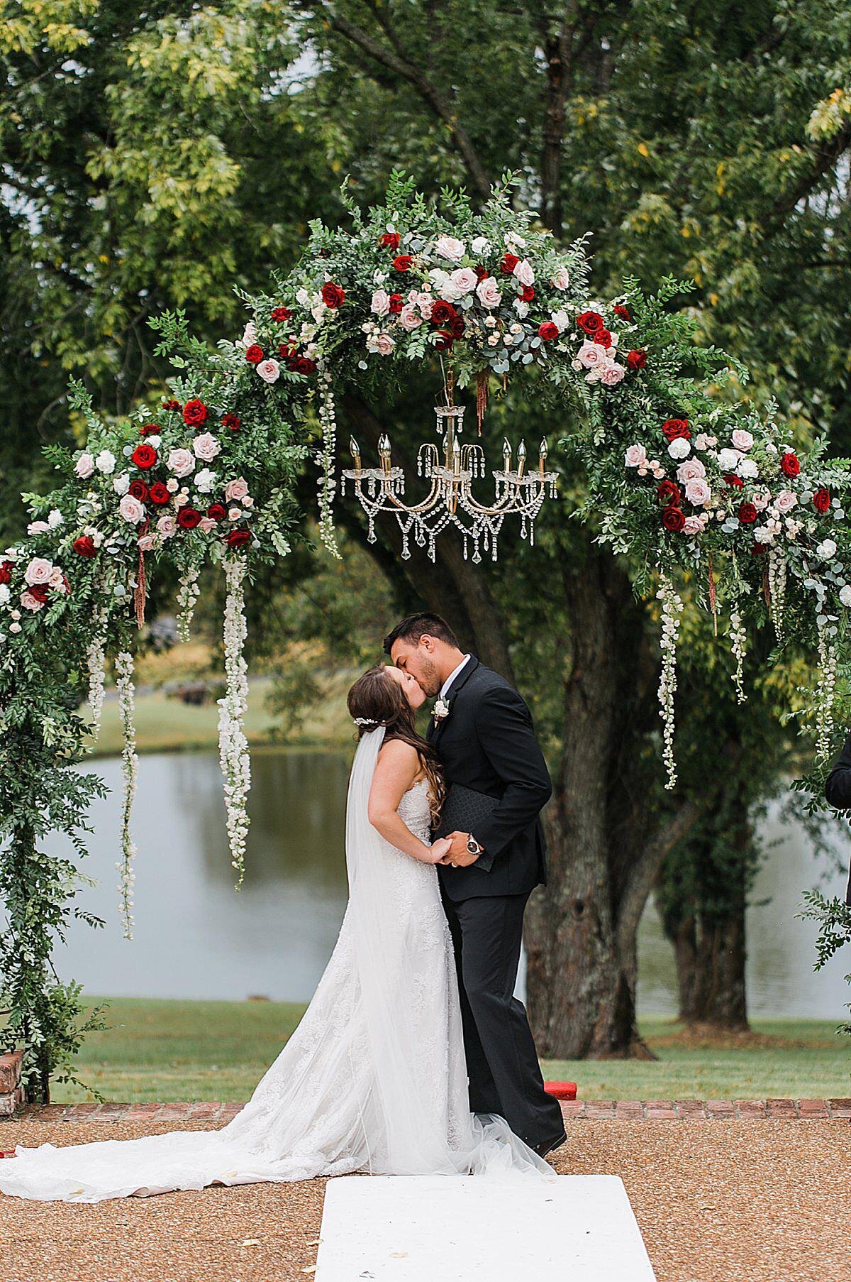 bride wearing a wedding dress with a long train and veil kisses the groom in a black tuxedo standing by a tree along a lake with an arbor of white, pink, blush and red roses and a crystal chandelier at their Nashville wedding
