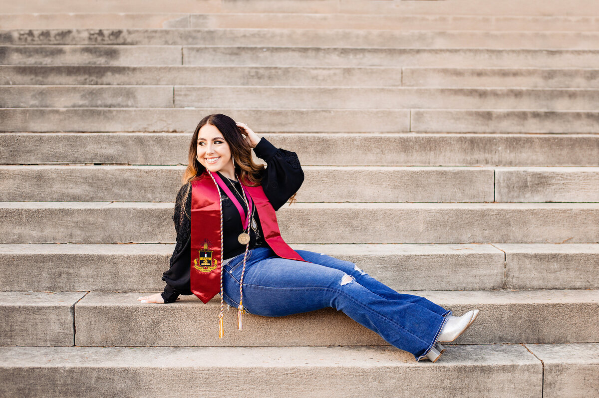 Graduation photo of girl wearing black shirt and jeans sitting on staircase wearing graduation cords