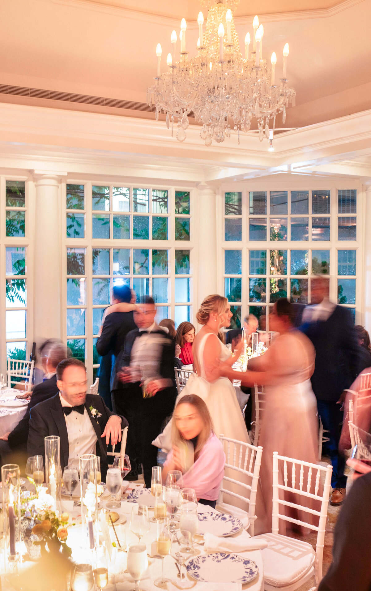 A vibrant wedding reception in a well-lit room with large windows. Guests in formal attire are mingling and chatting around elegantly set tables. A chandelier hangs from the ceiling, enhancing the festive atmosphere.