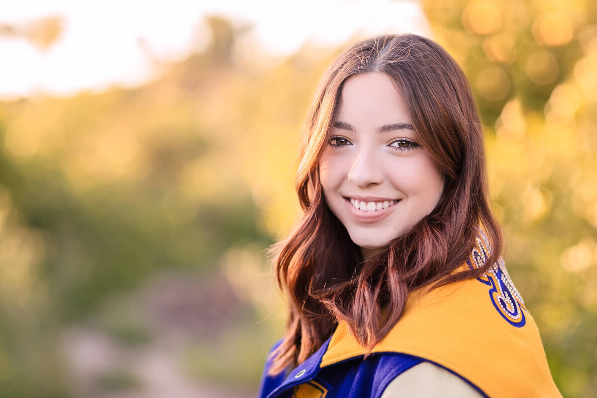 A close-up of a young woman with shoulder length, brown hair smiling at the camera and wearing a dark blue and yellow letterman jacket, with greenery in the background.