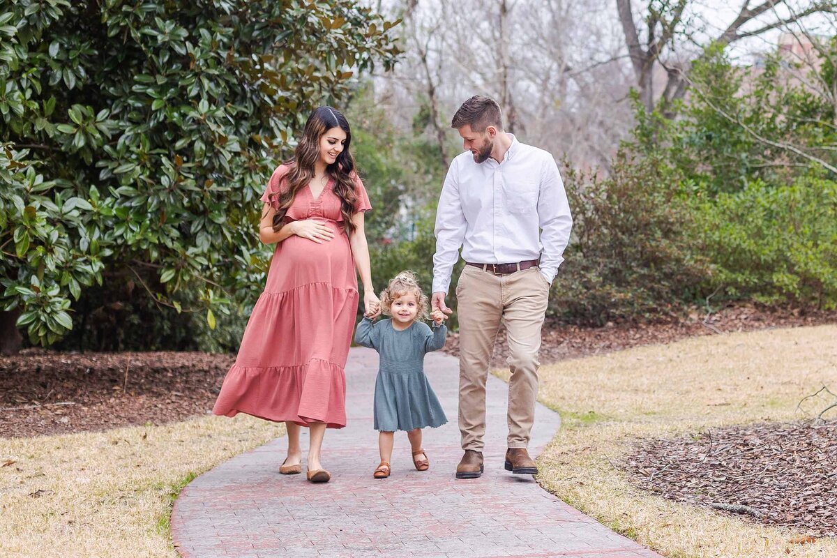 Family of three with a pregnant mom in a rose colored dress walking along a path at Palmetto Collegiate