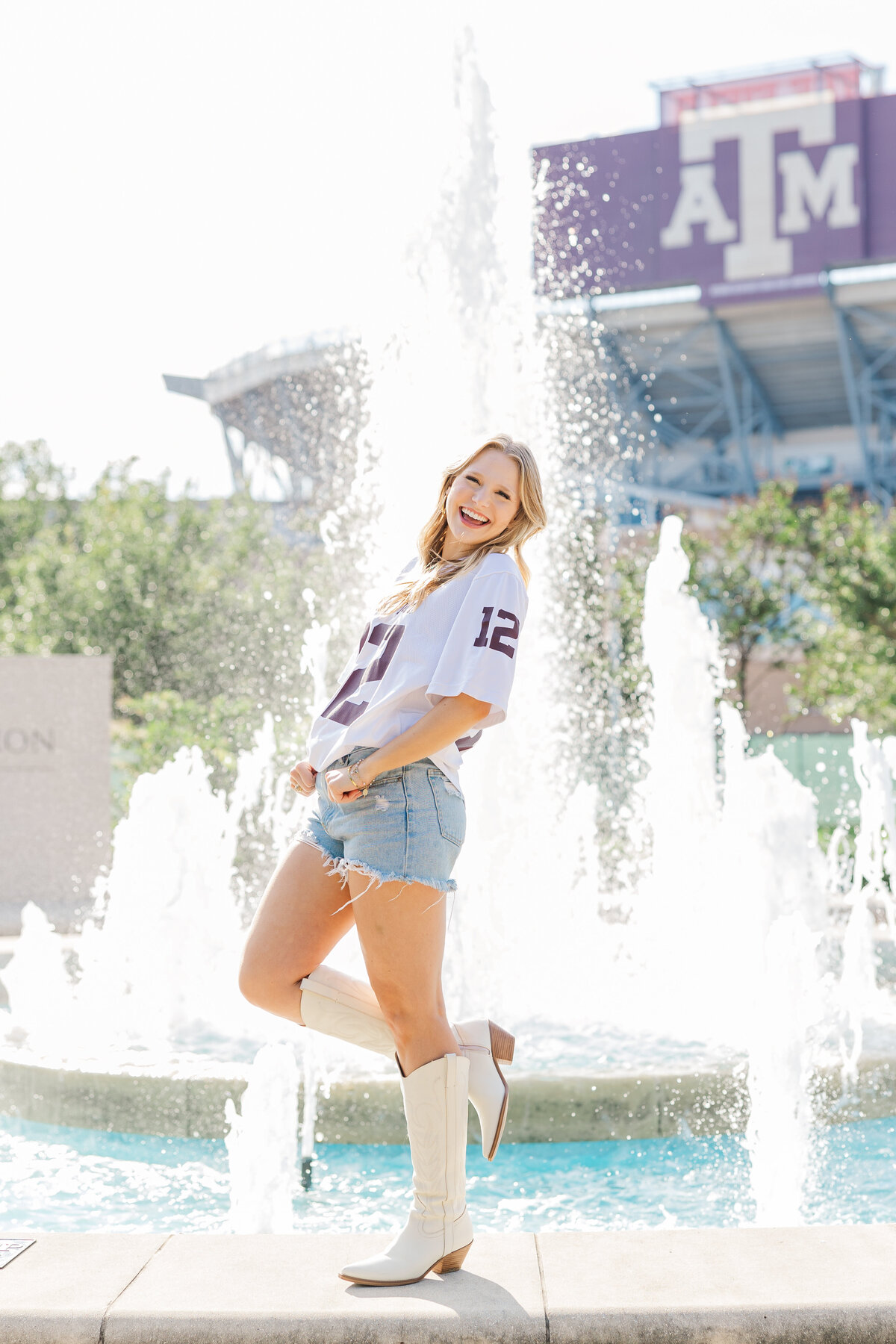 Smiling Texas A&M senior poses playfully in front of a sunny fountain wearing a 12th man jersey, denim shorts, and white cowboy boots. The stadium and large ATM logo are visible in the background, capturing school spirit and graduation joy in College Station.