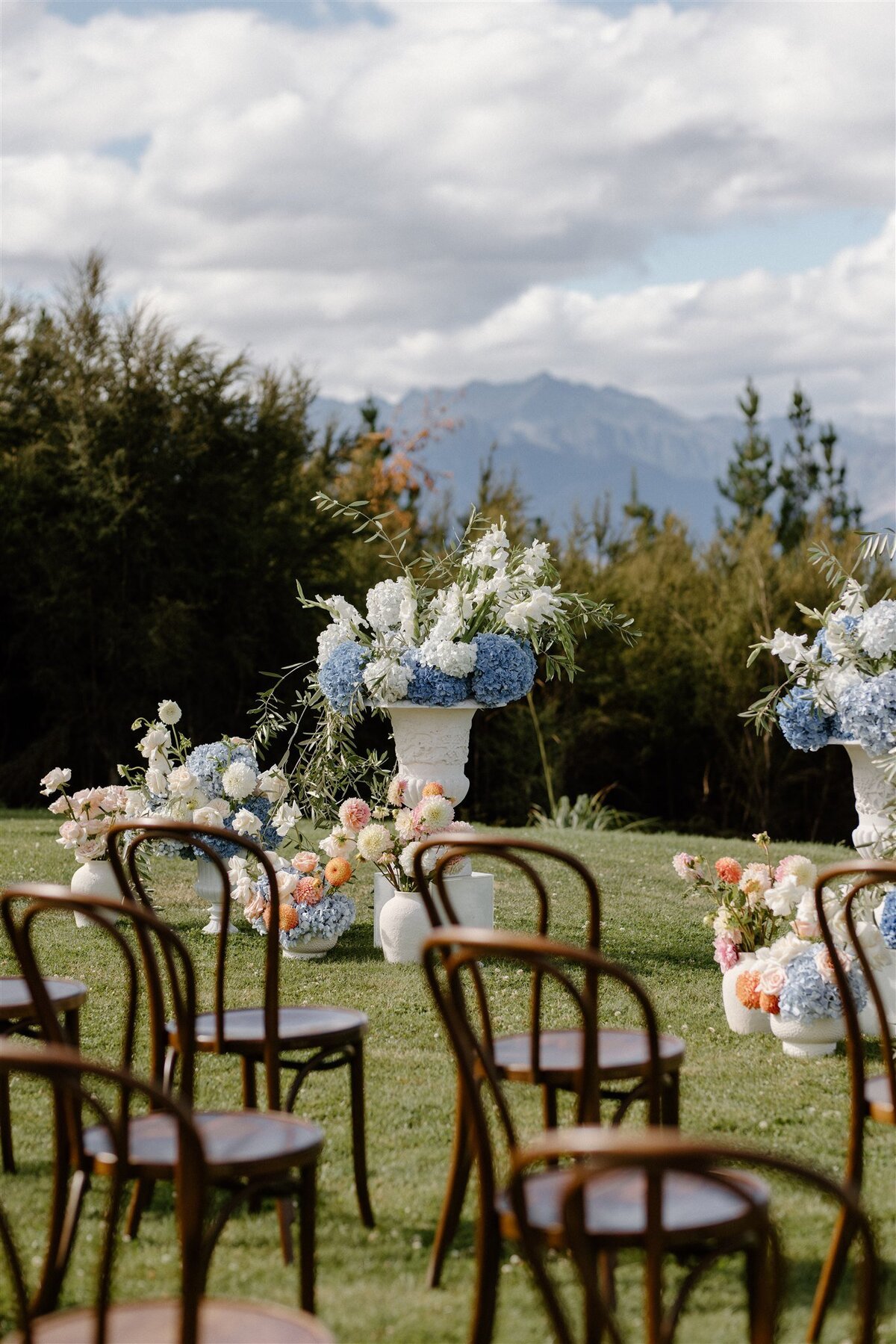 Lake Hāwea outdoor ceremony with rows of bentwood chairs and lush pastel flower arrangements in elegant white vases.