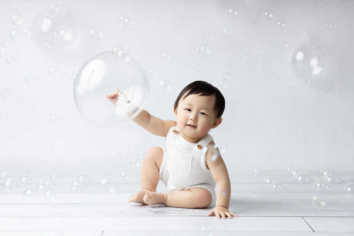 Dallas milestone photographer captures a happy baby in a white romper sitting surrounded by floating bubbles, playfully reaching out to pop one, with a joyful smile on their face.