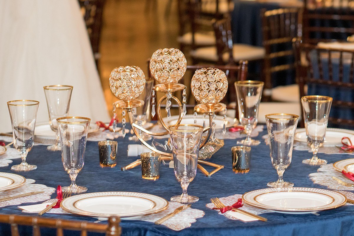 A gold and crystal candelabra sits on top of a table with a velvet navy table cloth. The table is set with footed crystal goblets with a gold rim, gold ginko leaf charger with a white and gold plate, matte gold flatware with a small thank you note tied to each fork and gold votive candles.