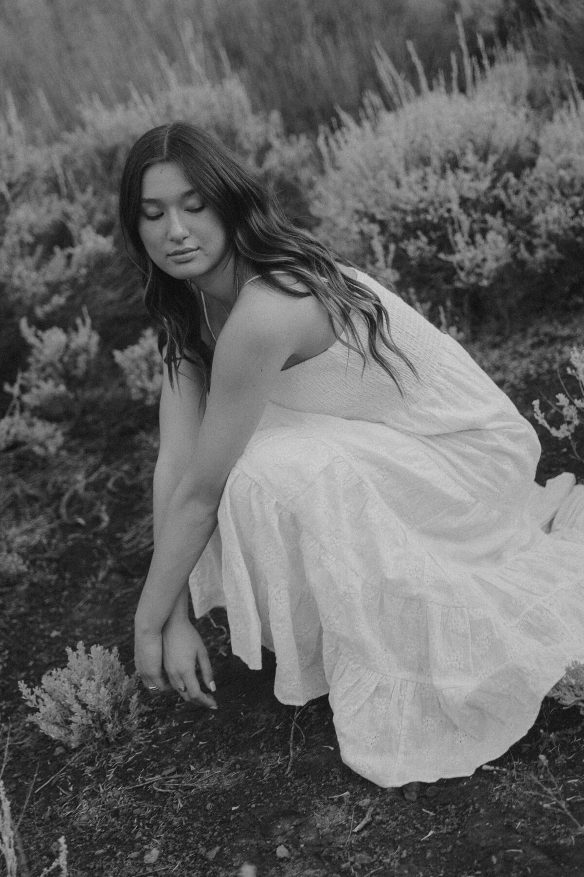 Black and White Senior Portrait of Girl Kneeling in Wild Sagebrush Field at Sunset