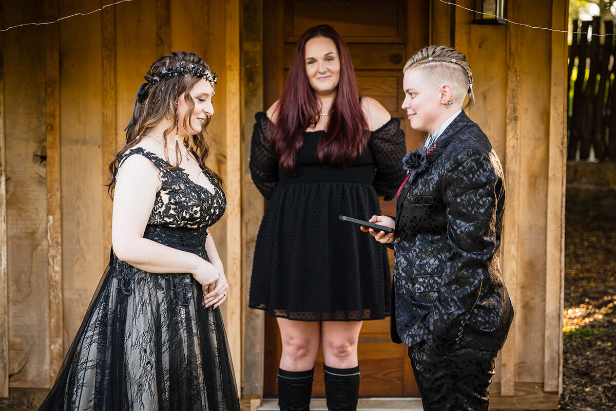 Two queer brides stand in front of one another, as one bride wearing a floral suit reads their vows written on their phone. A friend of theirs stands in the middle to act as their officiant for the day of their ceremony in a backyard of an Airbnb they rented.