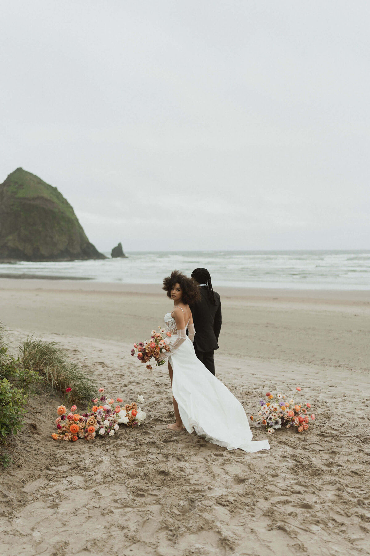 A candid wedding photo of a couple walking along Cannon Beach in Oregon. On the left is the bride holding a peach and purple wedding bouquet in her right hand and holding her partner's hand with her left. She is a black woman with shoulder length curly hair. She is wearing a white wedding dress. On the right, the groom is looking back at his partner smiling while holding her hand. He is a black man with longer braids. He is wearing a black suit with a white shirt.