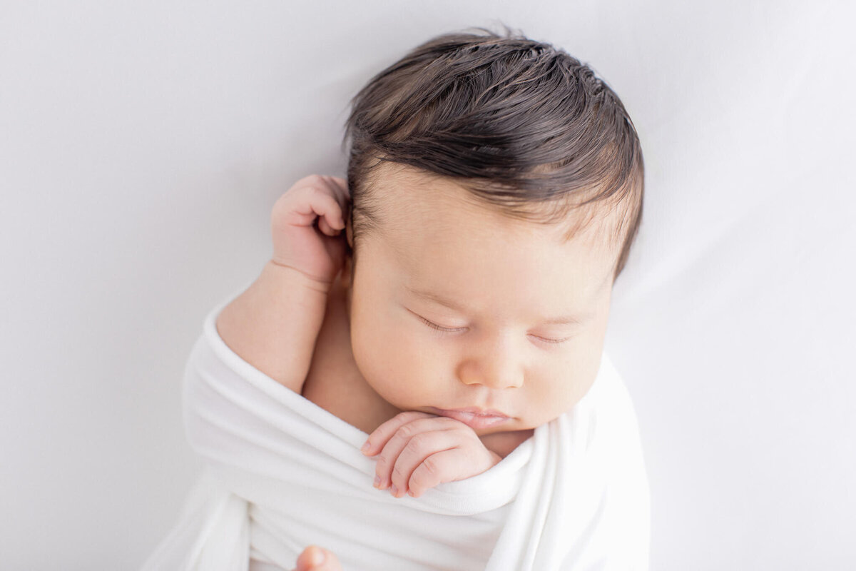 A sleeping baby wrapped in a white blanket rests peacefully on a soft, white surface. The babys dark hair is gently tousled, and one hand is tucked under their chin.