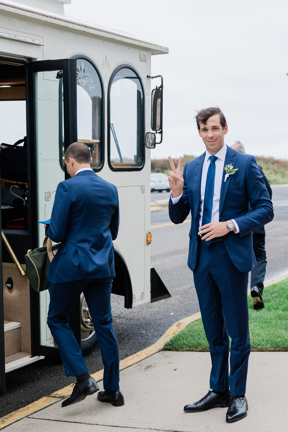 The groom, wearing a navy blue suit and boutonnière, flashes a peace sign while holding a drink as he stands outside a vintage white trolley