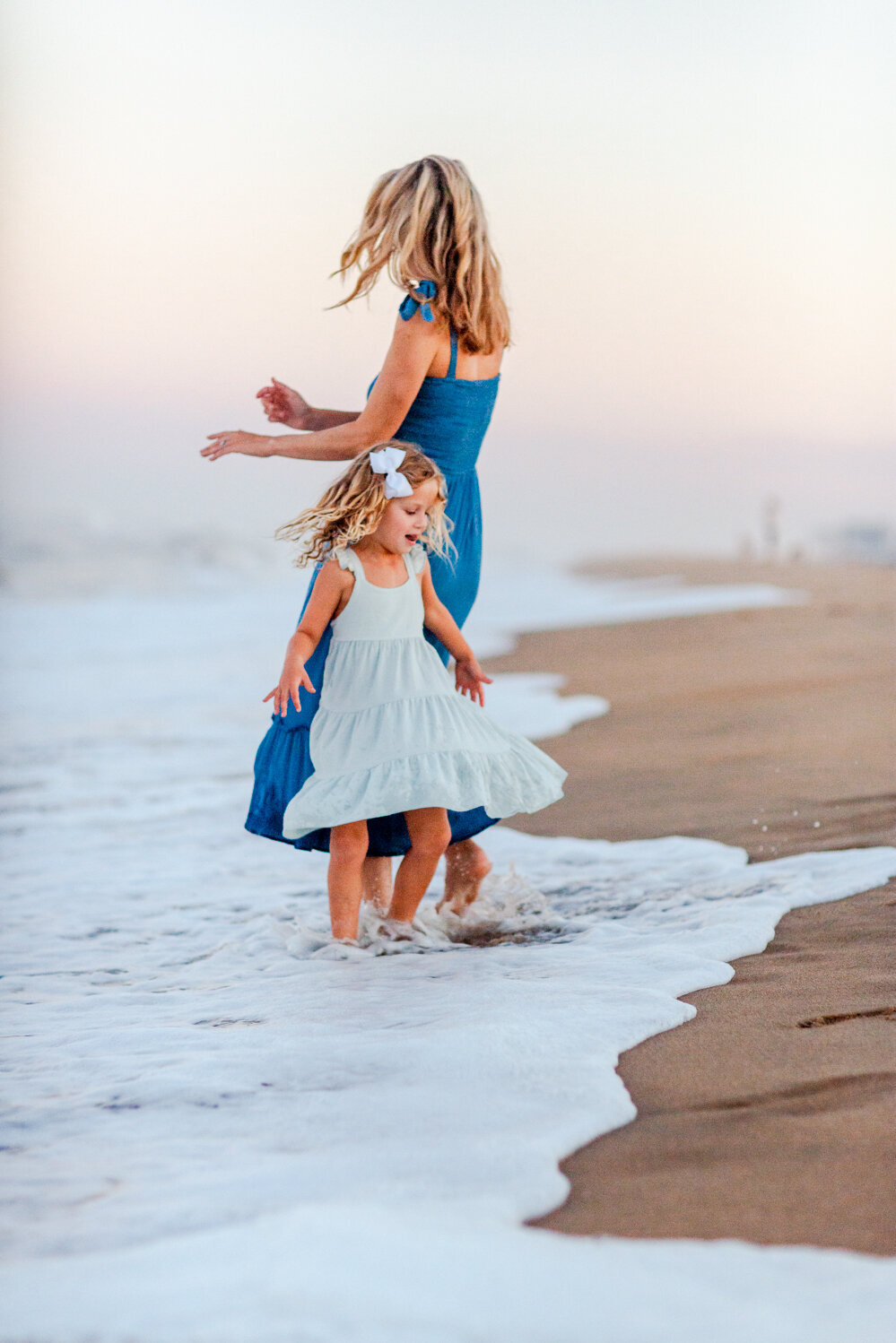 A joyful mother and daughter dance barefoot in the ocean waves at sunset, their dresses twirling as they together during a Coastal Virginia beach session with Brooke Tucker Photography