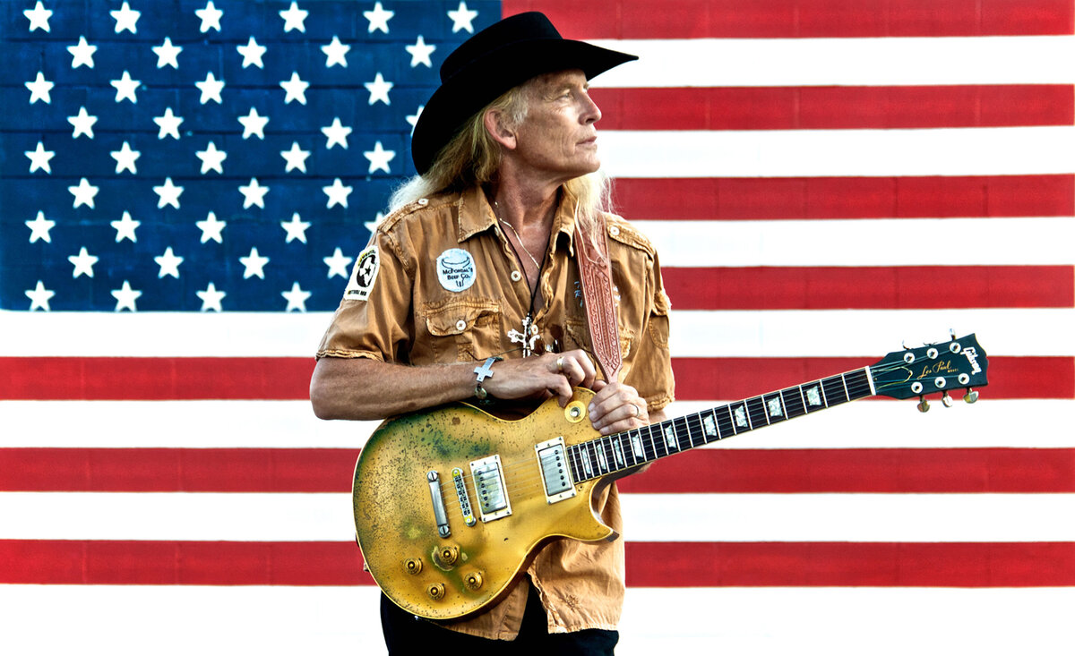 Country music photo Billy Crain holding gold electric guitar wearing black cowboy hat large American flag background