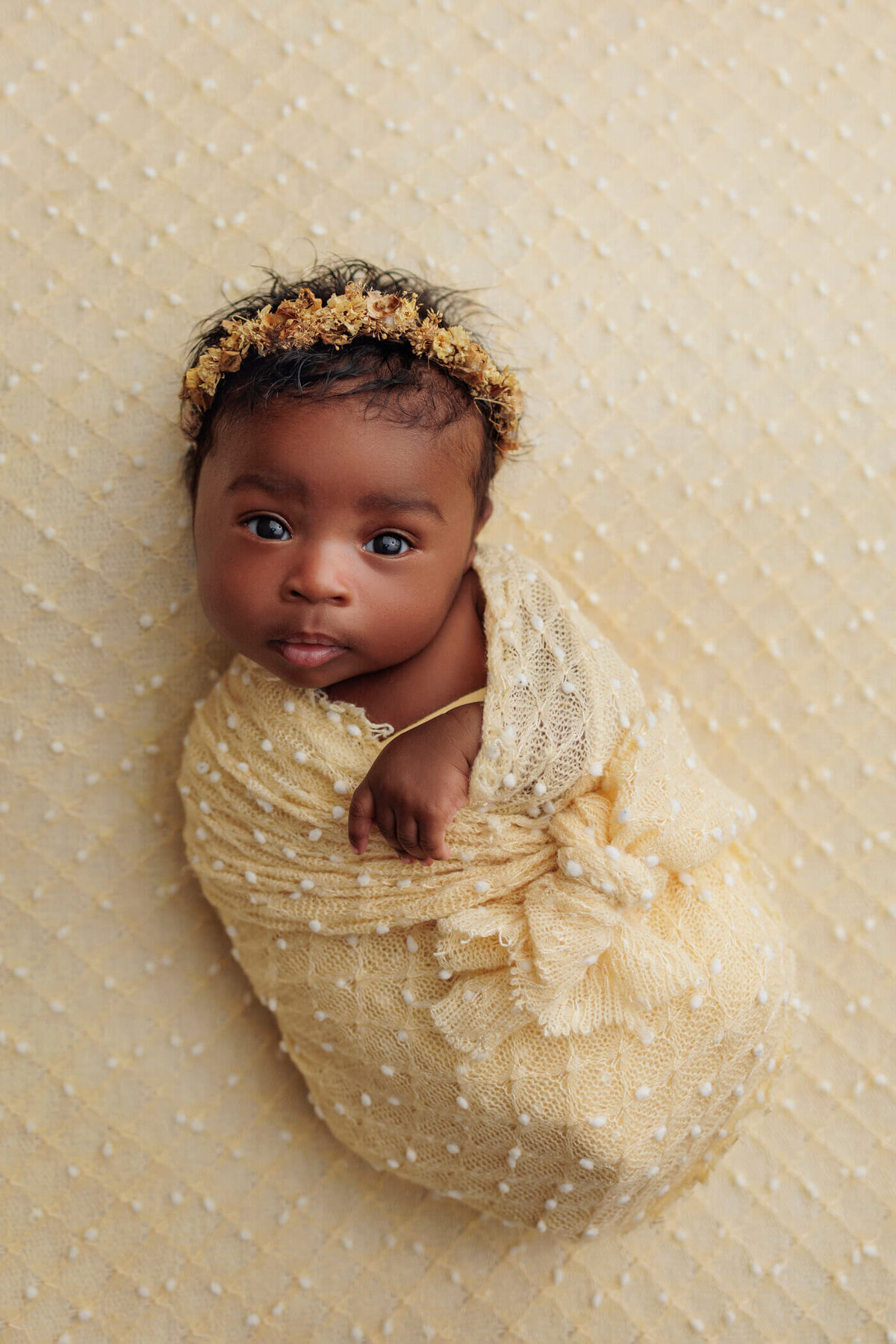 newborn baby girl wrapped in a soft yellow blanket, wearing a matching floral headband, lying on a yellow textured backdrop.