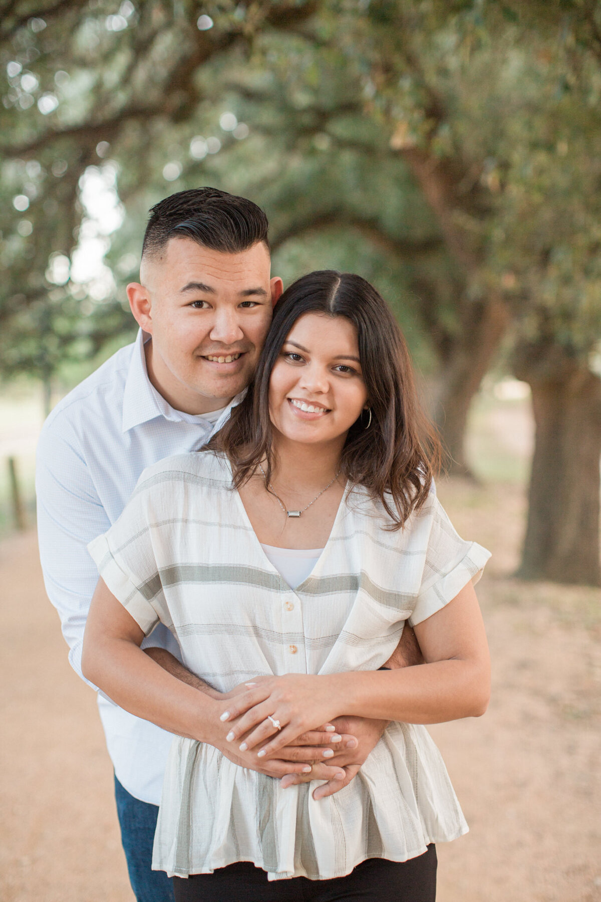 A man wraps his arms around his fiancee's waist and she rests her hands on his, displaying her ring while both smile at the camera