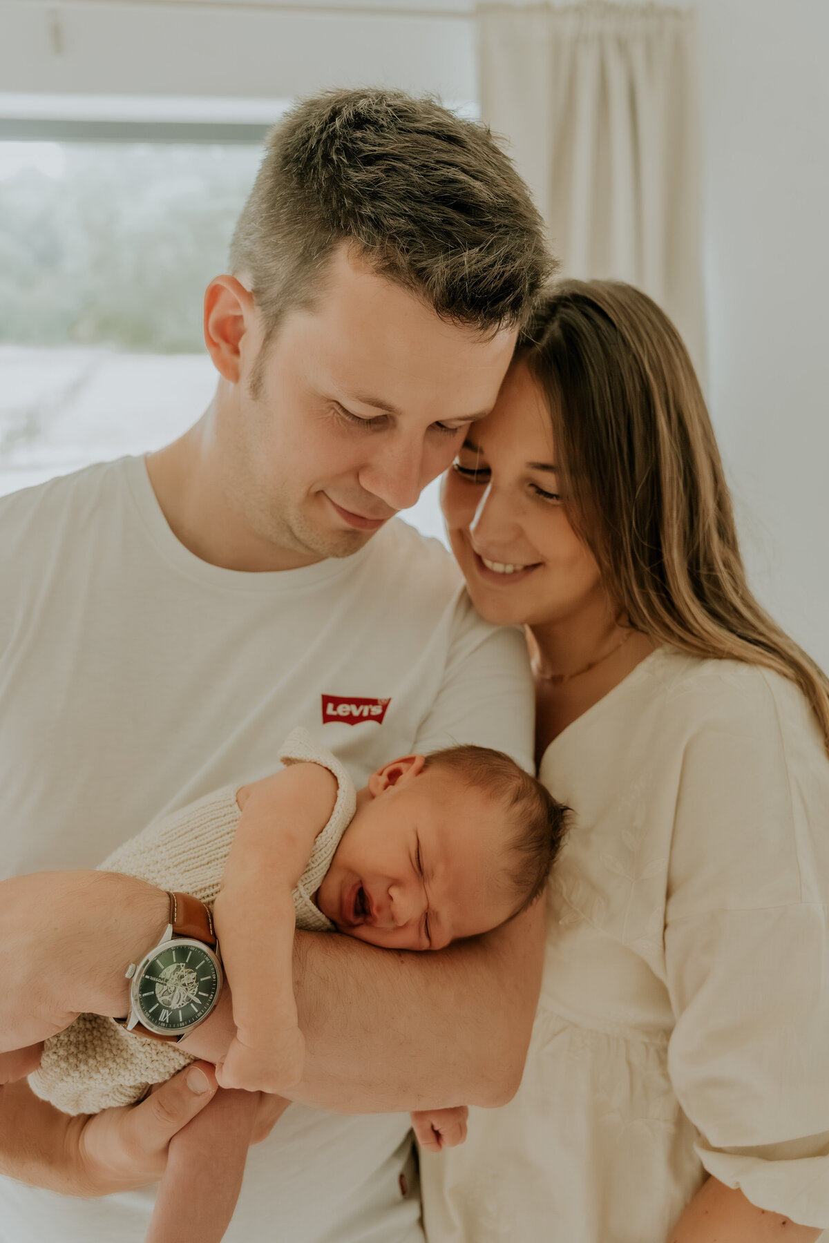 Newborn baby vredig in de armen van de papa, mama omhelst papa en baby en kijken beiden gelukkig toe. Newborn shoot aan huis te Antwerpen als een gezinsshoot en newborn lifestyleshoot.