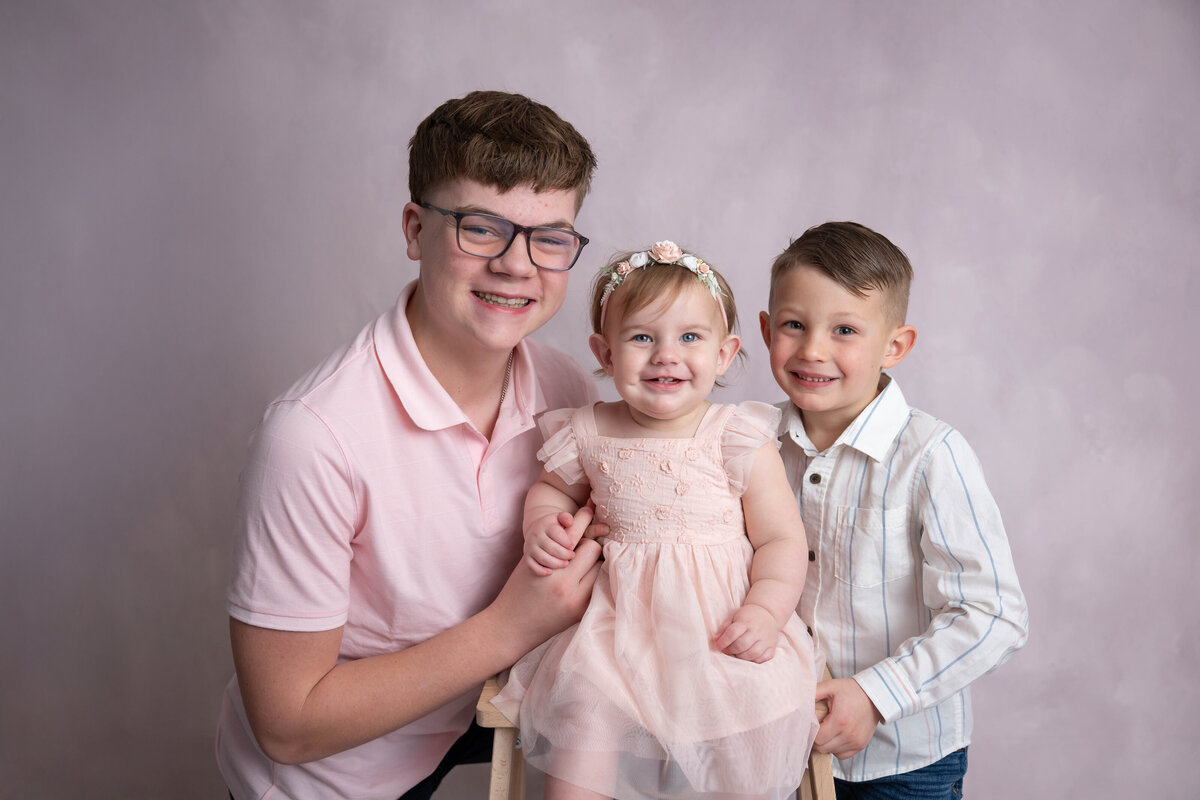 Toddler in a pink dress hugged on both sides by two older children, all wearing formal attire.