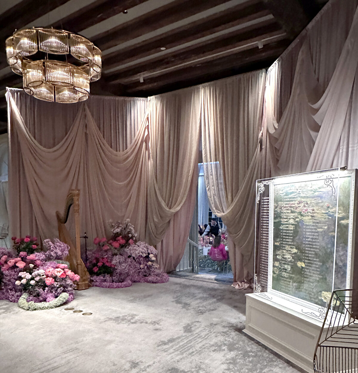 A luxurious wedding setup featuring rich pink draping, lush pink florals cascading to the ground, and an elegant glass case displaying guest table seating. The combination of soft textures, romantic hues, and modern sophistication creates a breathtaking entrance for an upscale wedding reception.