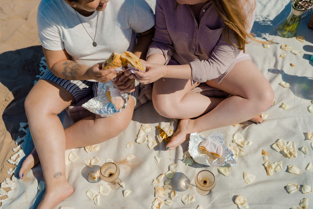 two women sitting on a blanket on the beach with champagne glasses and seashells on it