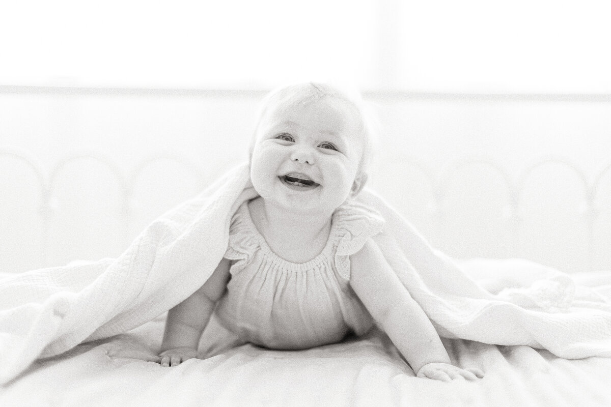 Black and white photo of a baby girl smiling and peeking out of a blanket as she smiles for her milestone session.