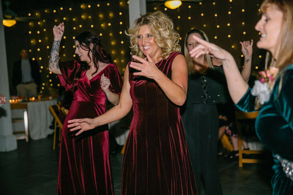 A group of women in formal dresses joyfully dancing, with string lights glowing in the background. The focus is on a smiling woman in a burgundy velvet dress in the center.