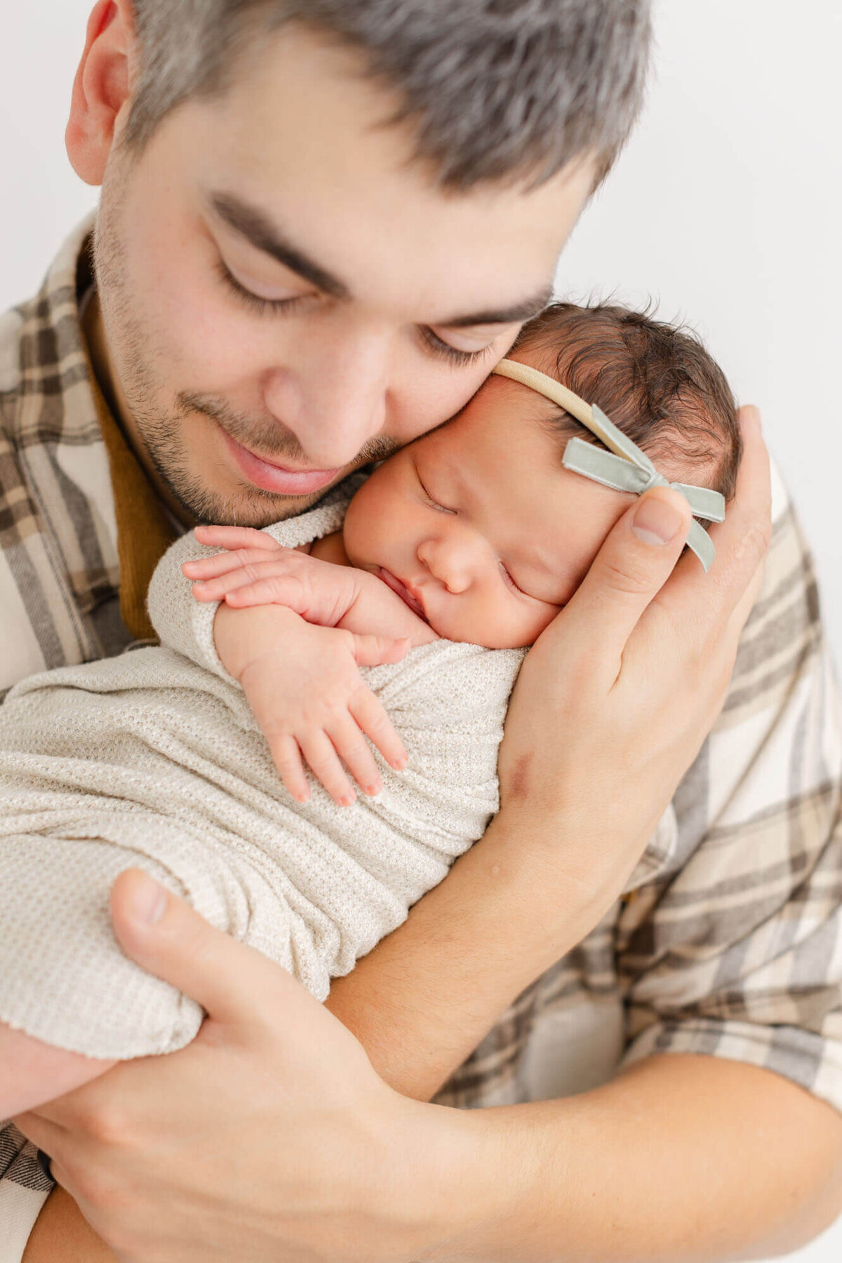 Dad holding newborn baby girl up to his cheek and his eyes are closed and he has a soft smile on his face. Dad and baby are both dressed in light colored neutrals.