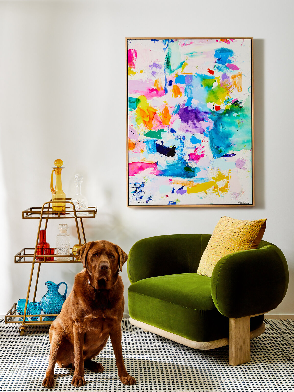 A green velvet armchair with a yellow pillow sits on a patterned rug in a Sandringham home. A brass bar cart and a brown Labrador dog are visible in front of a large, colorful abstract painting.