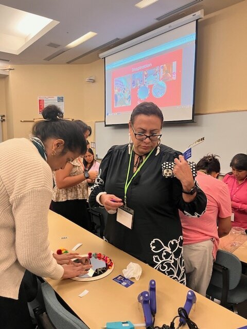 The creative journey as two women work on a wooden table, one carefully gluing vibrant pompoms around a round mirror while the other observes.