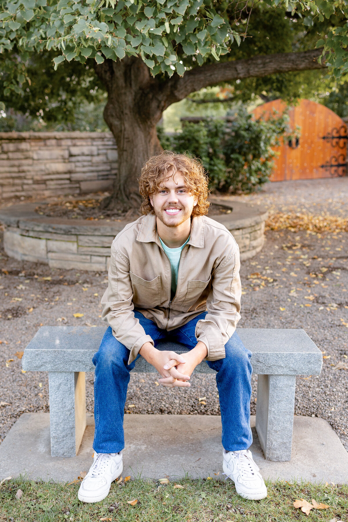 A young man with curly hair sits on a stone bench under a large tree, smiling at the camera. He wears a tan jacket and jeans, with a wooden gate and greenery in the background.