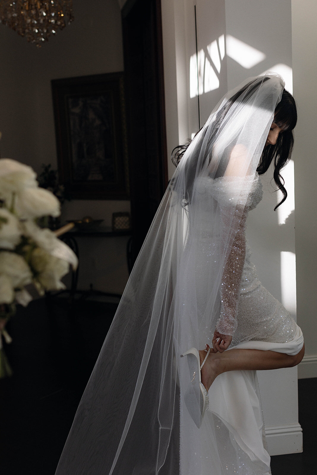 An artistic bridal portrait of a bride adjusting her gown, her veil cascading in soft light. A refined and editorial wedding photograph emphasizing elegance and candid beauty.