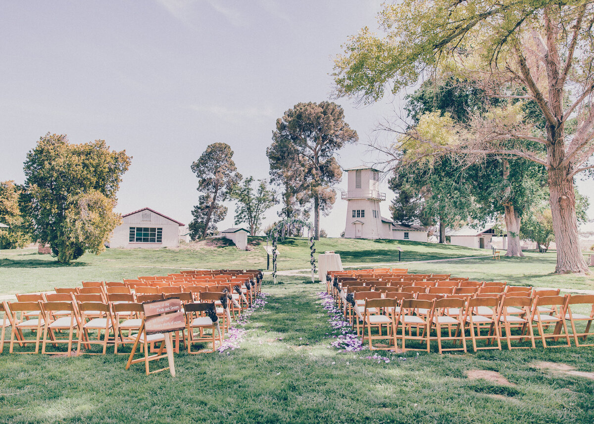 A rustic, simple outdoor wedding ceremony set with wooden chairs on green grass with trees and a barn building.