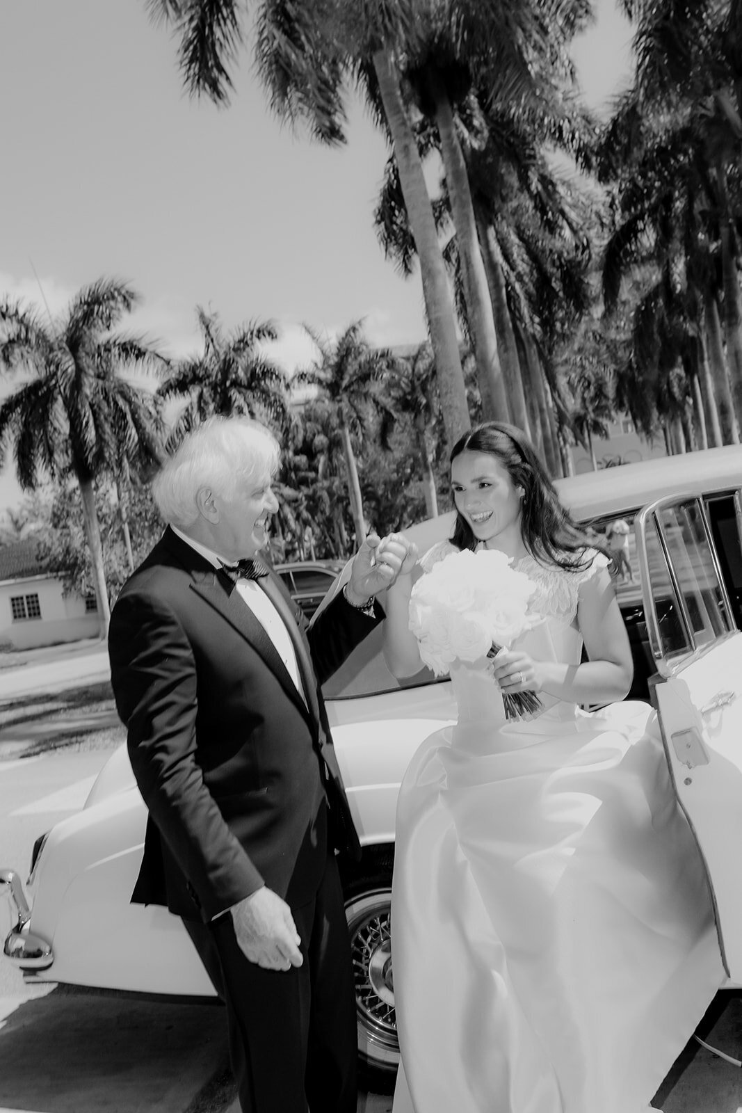 Black and white photo of father helping the bride out of the wedding car. He smiles at her and she smiles back at him.