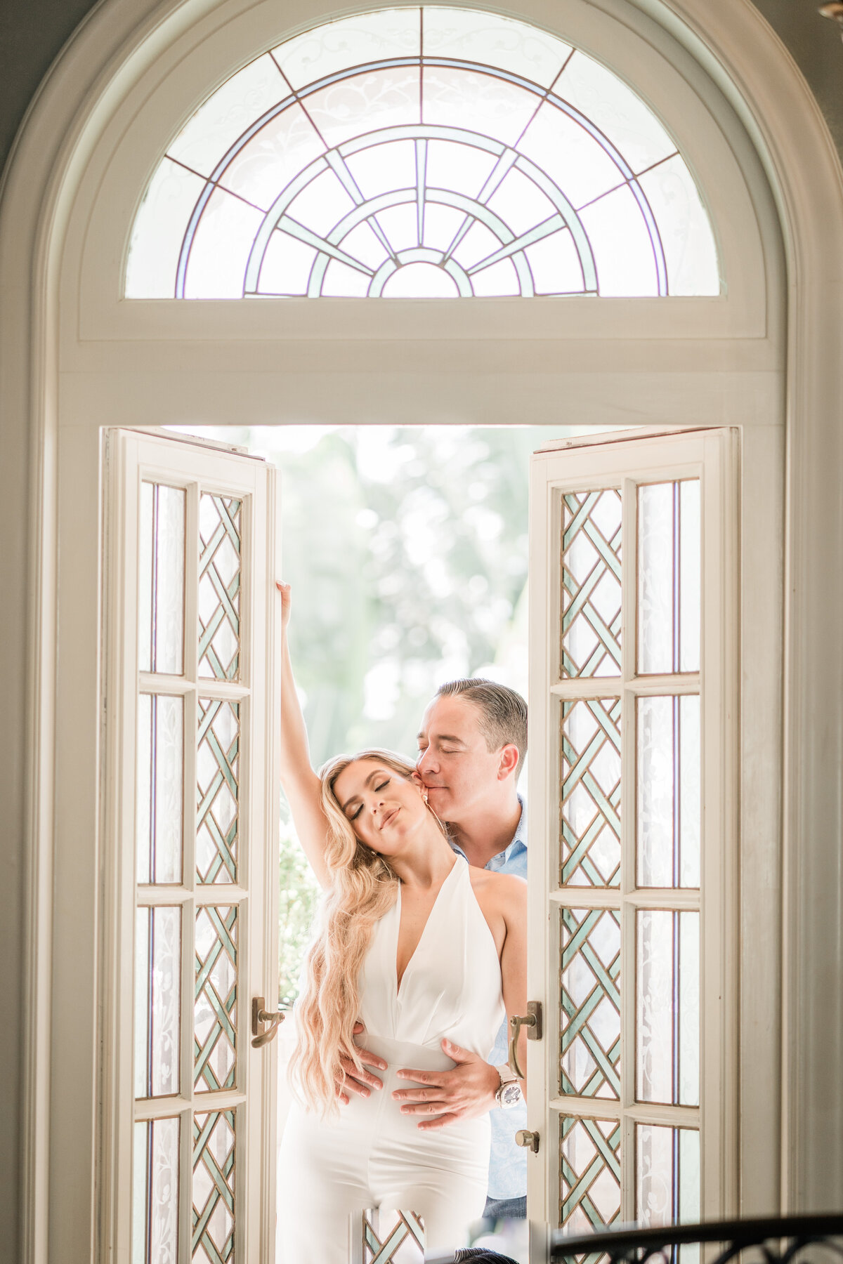 Romantic engagement portrait of a couple embracing by elegant French doors in Miami, Florida, captured by Rodrigo Varela Photography. Intimate and timeless setting for a love-filled moment.