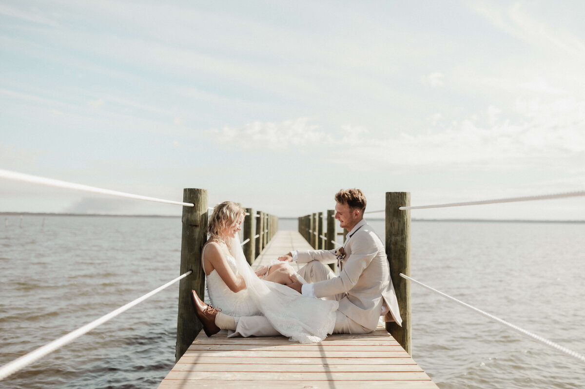 young newlywed couple sits on pier, holding hands after intimate wedding photographed by Brittney Stanley of Be Seen Photos