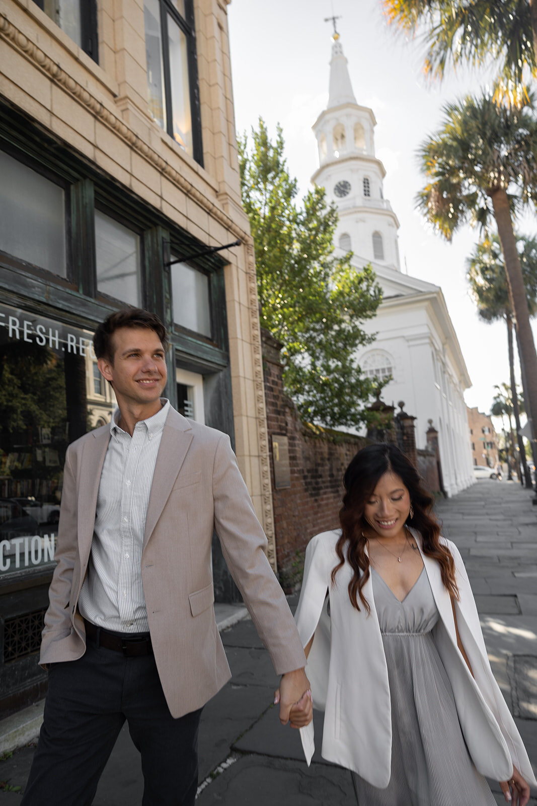 Couple walking down boardwalk on Braod Street in Charleston. St Michaels Church in background