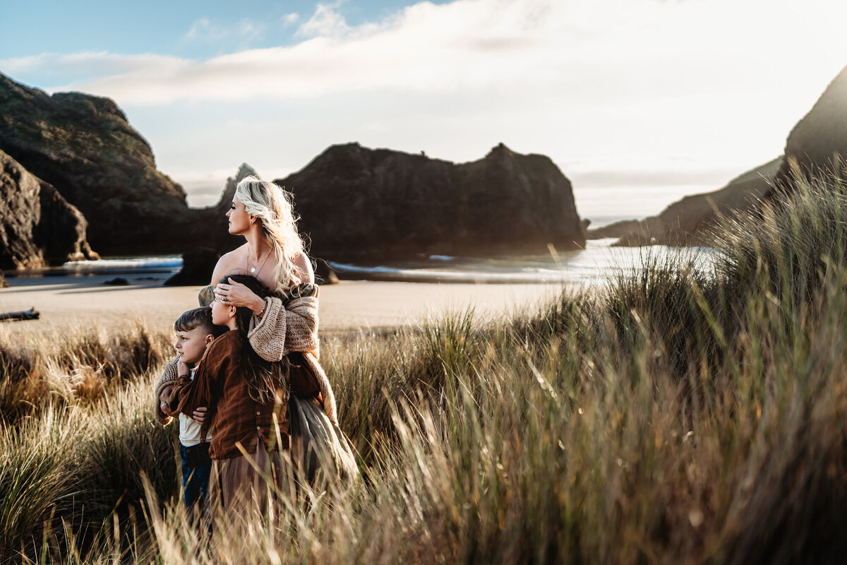 A mother holds her two children as they watch the sunset on the souther Oregon coast, during a lifestyle family photography session by travel family photographer Love Michelle Photography