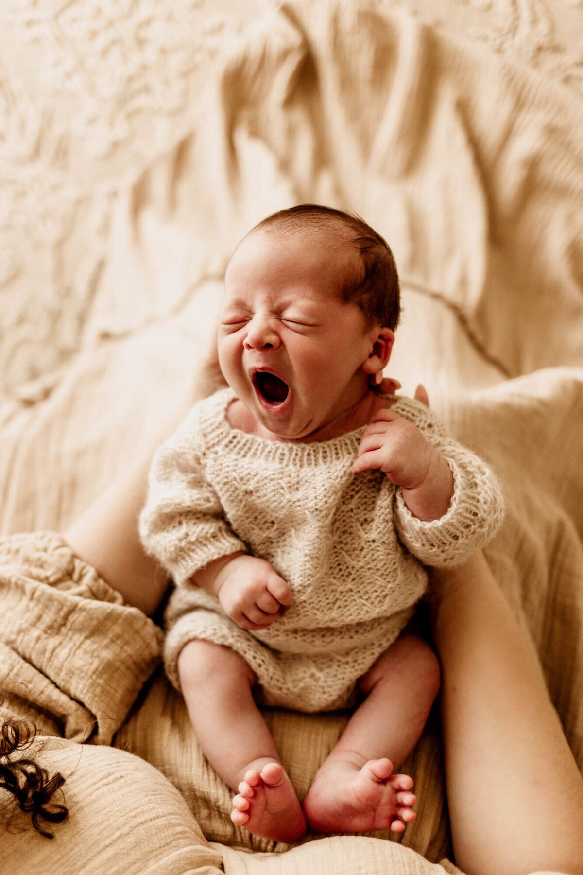 Baby boy wearing a hand knit beige romper held in his mother's arms yawning.