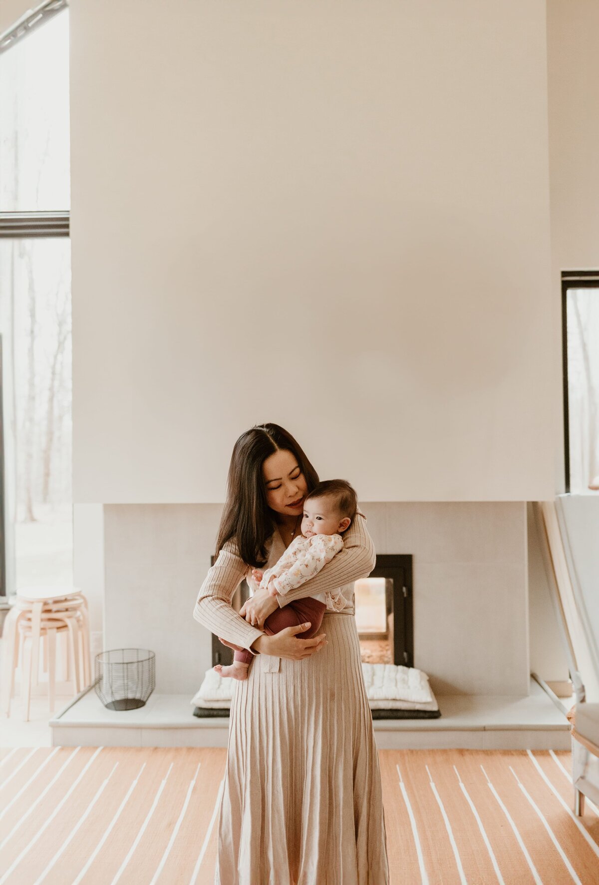 A mother lovingly cradles her baby in a modern living room with large windows, a fireplace, and chic minimalist decor.